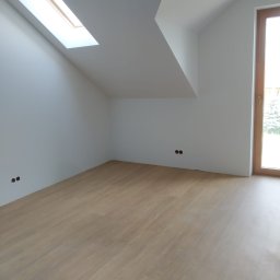 Unisarwan Szczupakowska Anna - Poddasze ze skośnym dachem i oknem dachowym, świeżo wykończone gładzią, z jasną drewnianą podłogą i widocznymi puszkami elektrycznymi w ścianie, gotowe do dalszych prac wykończeniowych.