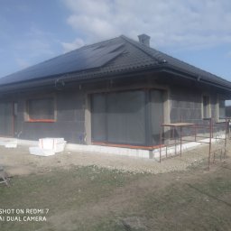 Unisarwan Szczupakowska Anna - Surowy stan domu jednorodzinnego z szarego bloczka, przygotowanego do ocieplenia, z zamontowanymi panelami słonecznymi na dachu i folią zabezpieczającą okna, widoczne rusztowanie i pracownik przy...