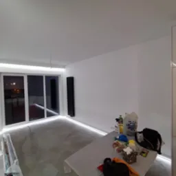 Pokój w trakcie remontu z oświetleniem LED przy suficie i podłodze, widoczne narzędzia i materiały budowlane na stole, szare ściany i posadzka, czarny grzejnik na ścianie.