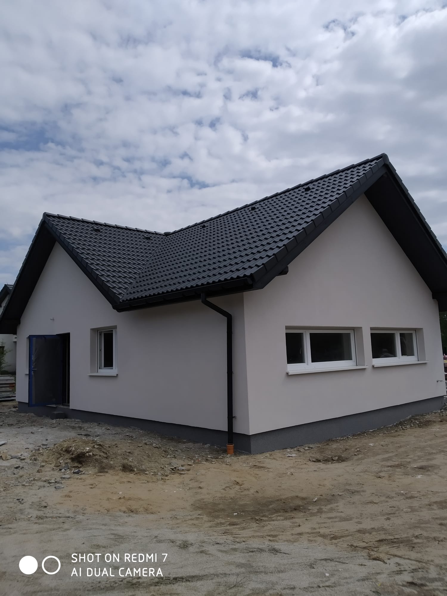 Nowo wybudowany dom jednorodzinny z grafitową dachówką i jasną elewacją w trakcie wykańczania terenu wokół budynku. Widoczne nieotynkowane fragmenty elewacji i otwarte drzwi wejściowe.