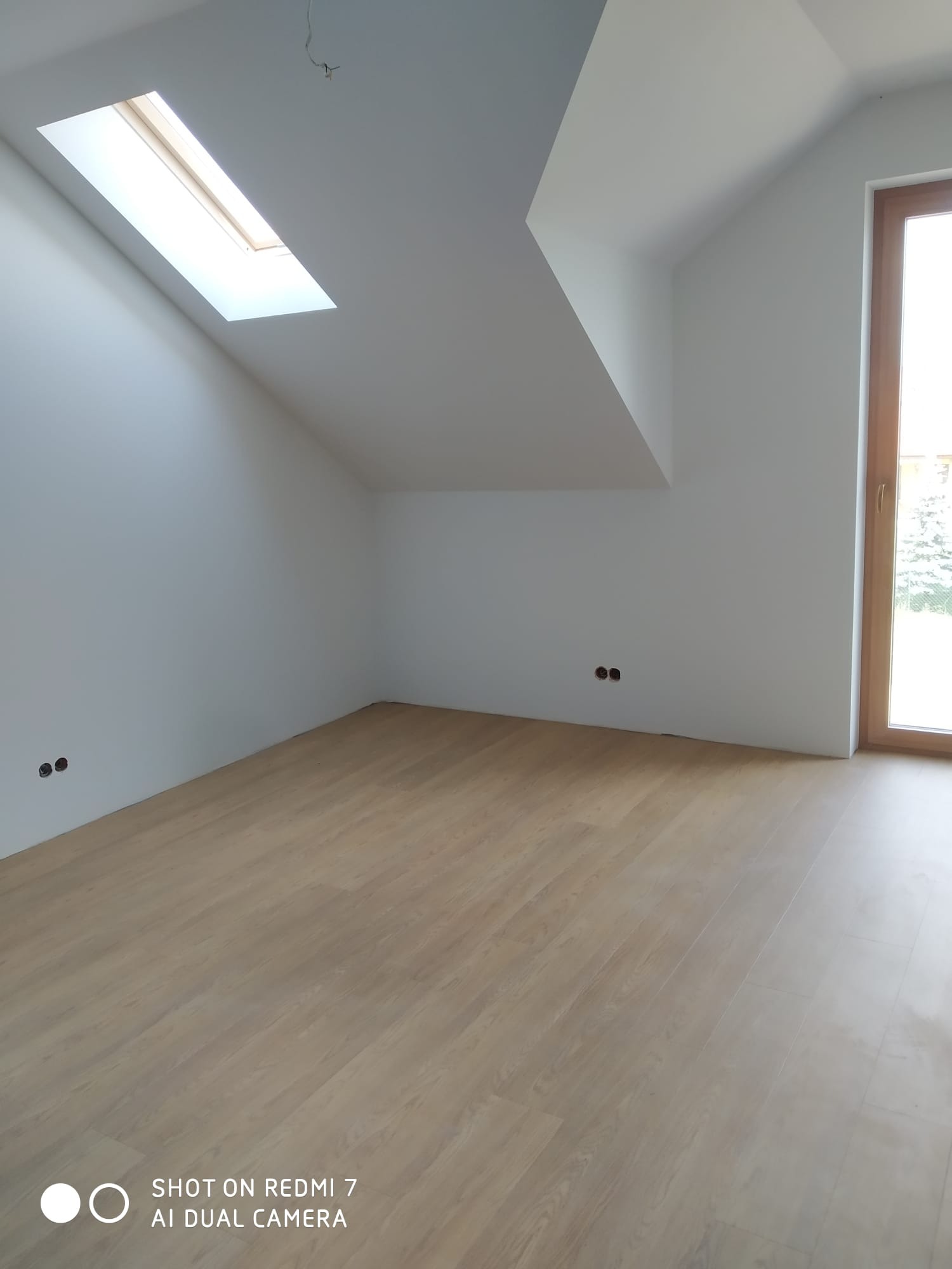 Poddasze ze skośnym dachem i oknem dachowym, świeżo wykończone gładzią, z jasną drewnianą podłogą i widocznymi puszkami elektrycznymi w ścianie, gotowe do dalszych prac wykończeniowych.