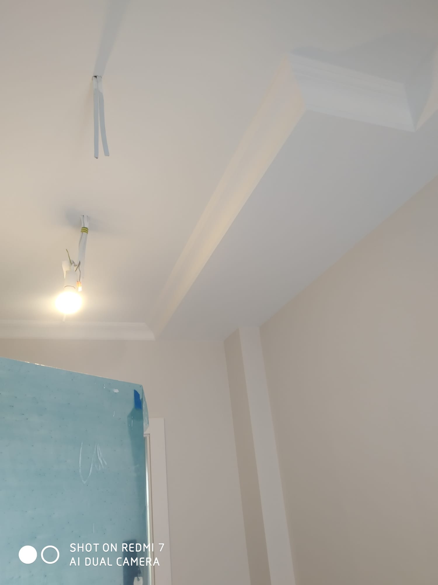 Szczegółowe ujęcie wykończonego sufitu z dekoracyjnymi elementami i wystającymi kablami elektrycznymi, oświetlonego żarówką, z fragmentem zabezpieczonej folią ściany w dolnym rogu.