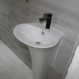 Biała umywalka na postumencie z czarną baterią, zamontowana przy ścianie wyłożonej płytkami imitującymi drewno, z widocznym surowym wykończeniem ściany po prawej stronie.