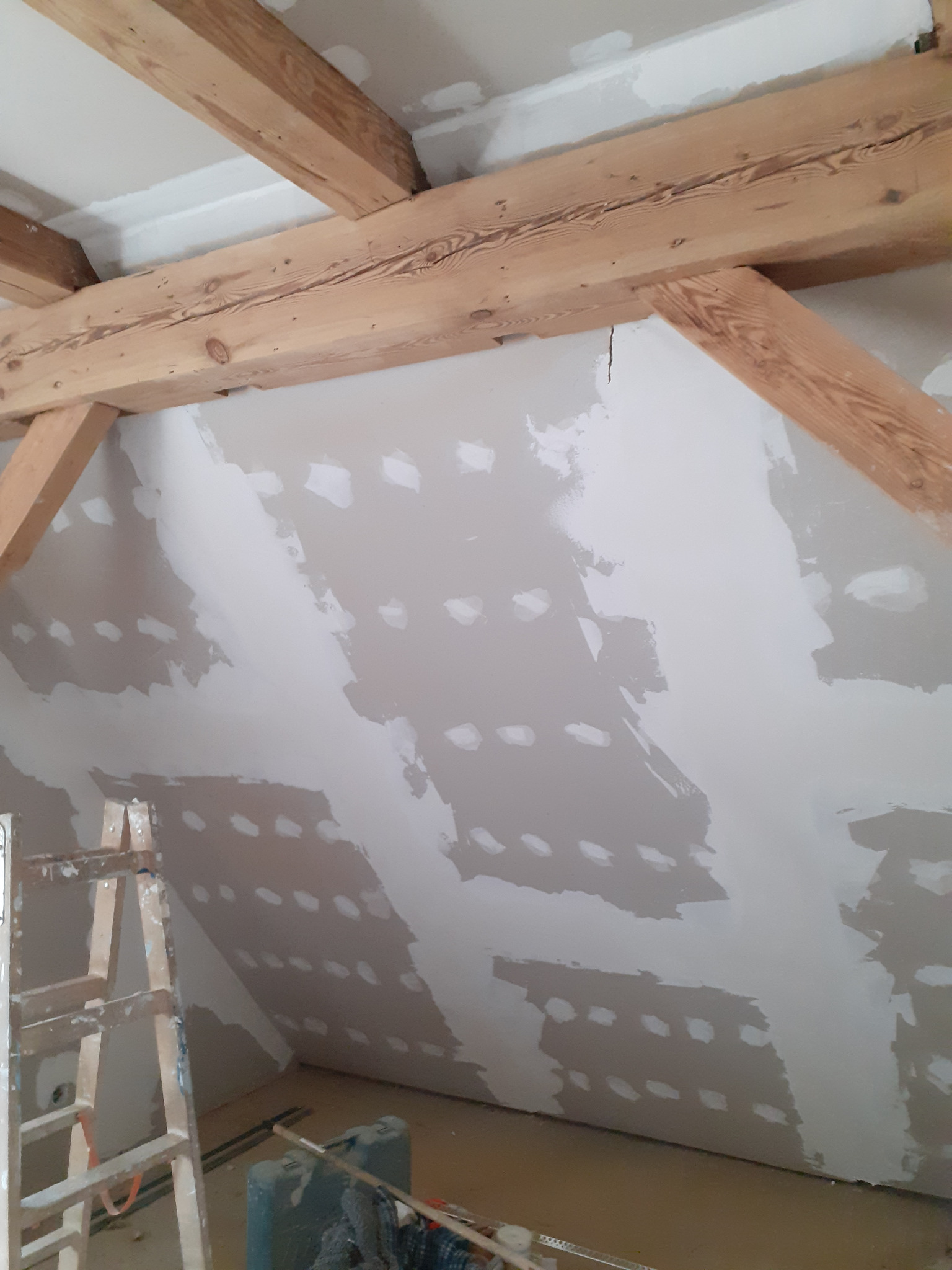 Poddasze w trakcie prac wykończeniowych: drewniane belki stropowe, ściana ze śladami po szpachlowaniu i nakładaniu gładzi, drabina malarska i akcesoria budowlane w pomieszczeniu.
