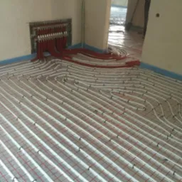 Białe rury ogrzewania podłogowego ułożone na czerwonej siatce, z czerwonymi rurami doprowadzającymi do rozdzielacza na ścianie, przed wylaniem posadzki.