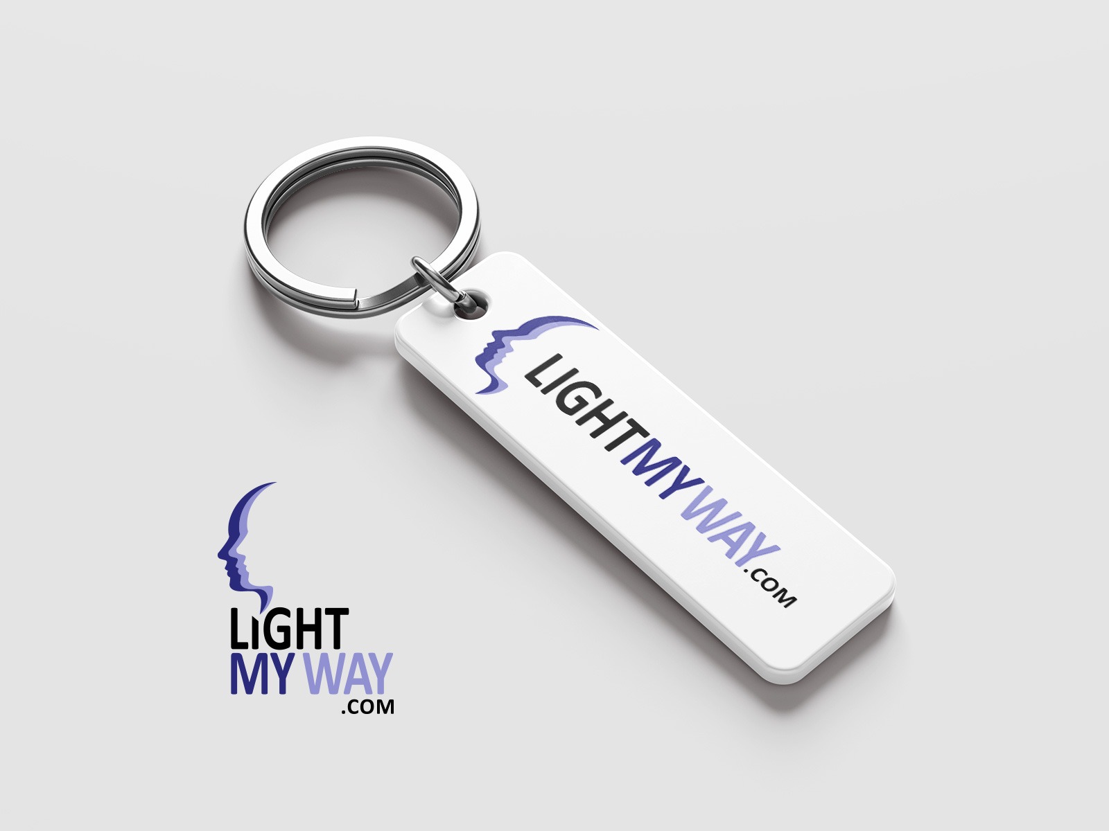 Biały brelok z logo 'LIGHTMYWAY.com' i profilem twarzy w odcieniach fioletu, leżący na jasnoszarym tle, obok wydrukowane logo firmy.
