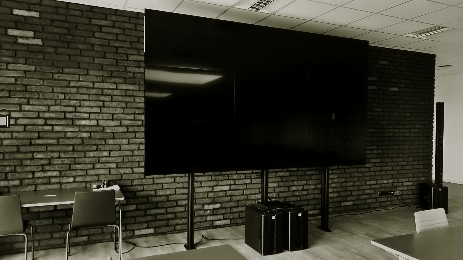 Duży, czarny ekran podzielony na trzy części, zamontowany na metalowych nogach w pomieszczeniu z ceglaną ścianą; widoczne głośniki kolumnowe i stoliki