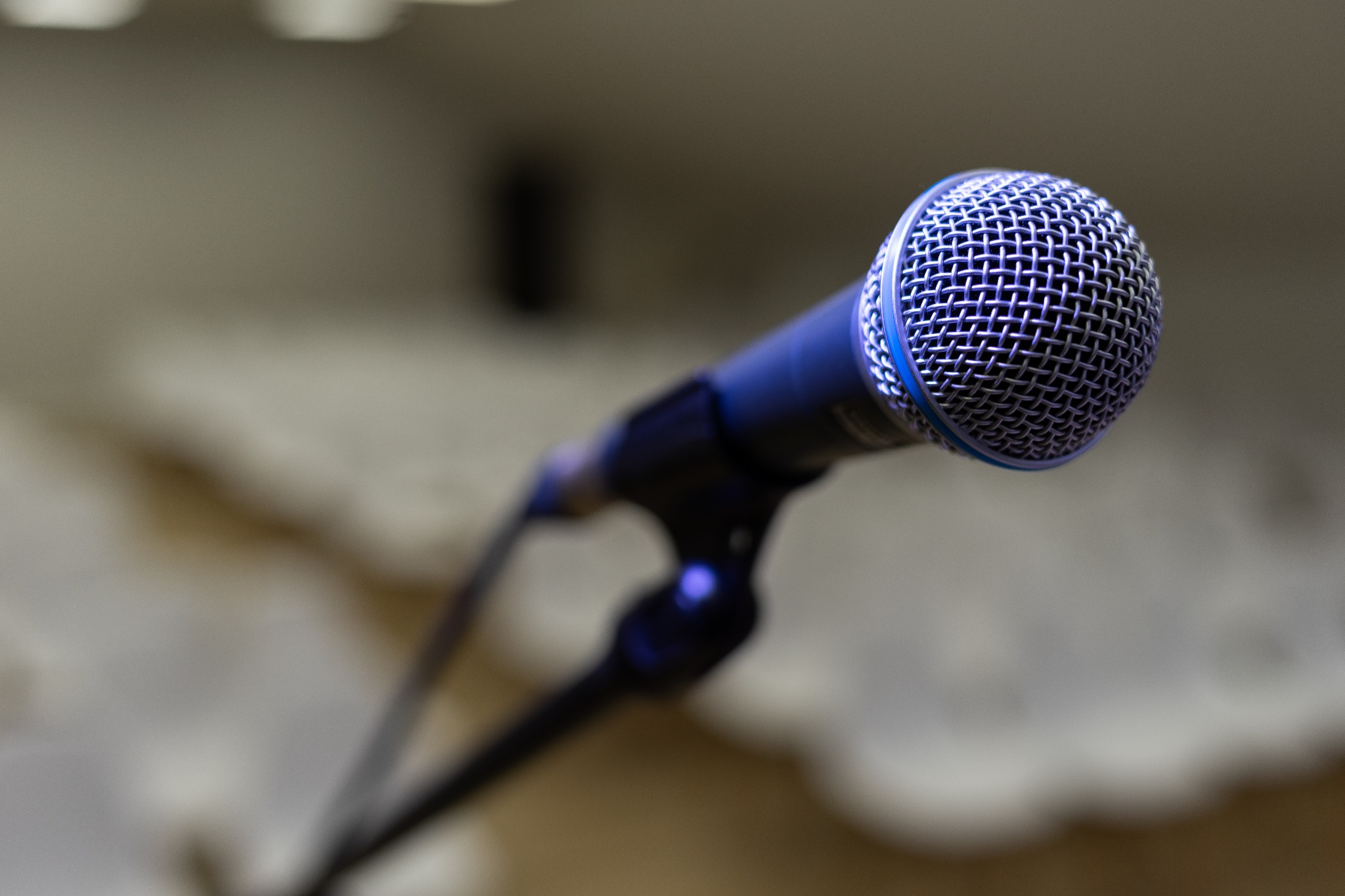 Zbliżenie na niebieski mikrofon dynamiczny na statywie, z metalową siatką ochronną na tle jasnej, rozmytej ściany. Widoczne detale konstrukcji mikrofonu i statywu.
