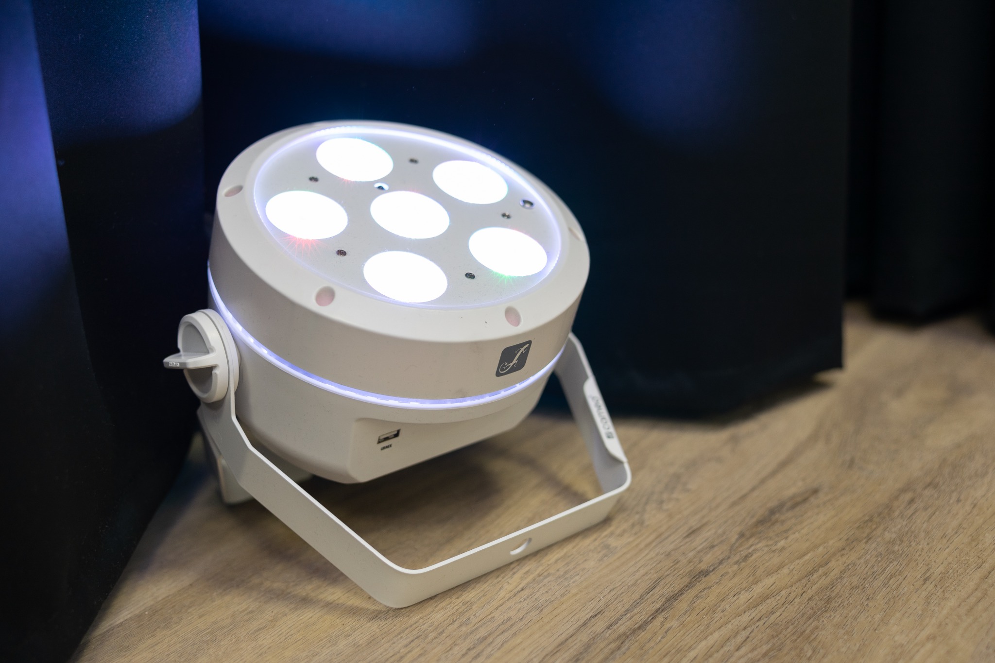Biała lampa LED PAR z sześcioma soczewkami, oświetlająca scenę, widoczne porty USB i DMX, obudowa z logo producenta, ustawiona na drewnianej powierzchni.