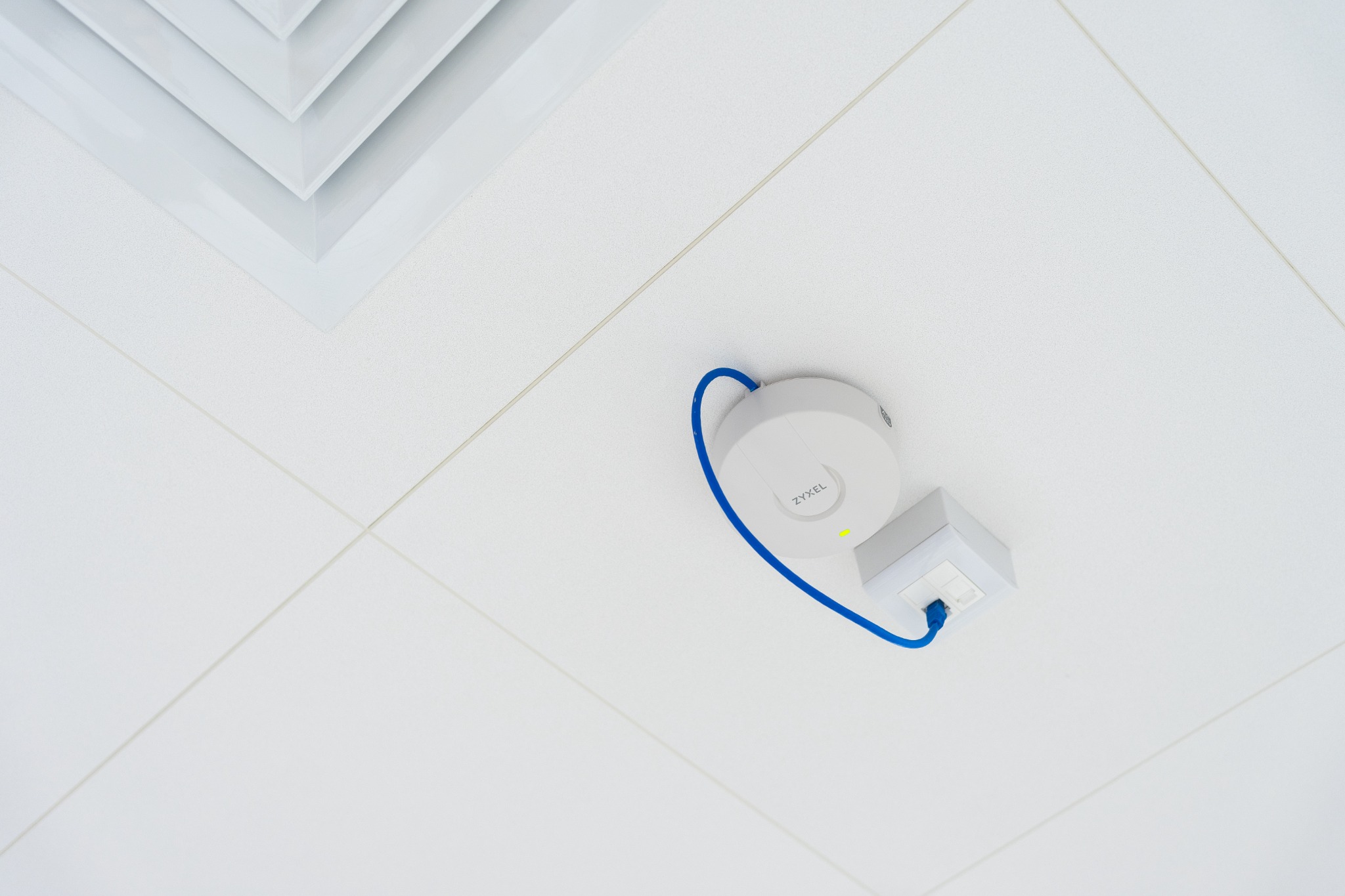 Biały punkt dostępowy Zyxel zamontowany na białym suficie podwieszanym z widoczną kratką wentylacyjną, podłączony niebieskim kablem ethernet do gniazda w ścianie.