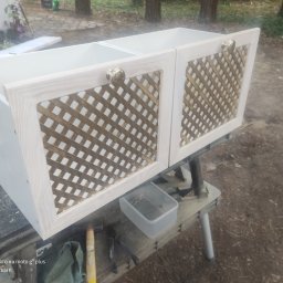 Usługi stolarskie - Dwa białe fronty szafek z ozdobną, ażurową kratką w kolorze złota i okrągłymi, metalowymi uchwytami, ustawione na warsztatowym stole, widoczne narzędzia i pudełko ze śrubami.