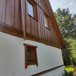 Usługi stolarskie - Fragment ściany domu z drewnianą elewacją i oknami z drewnianymi ramami, widok z dołu na tle zieleni.