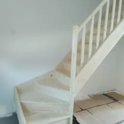 Surowe, sosnowe schody wewnętrzne z balustradą, widoczne zabezpieczenia podłogi kartonem i folią, świeżo po montażu.