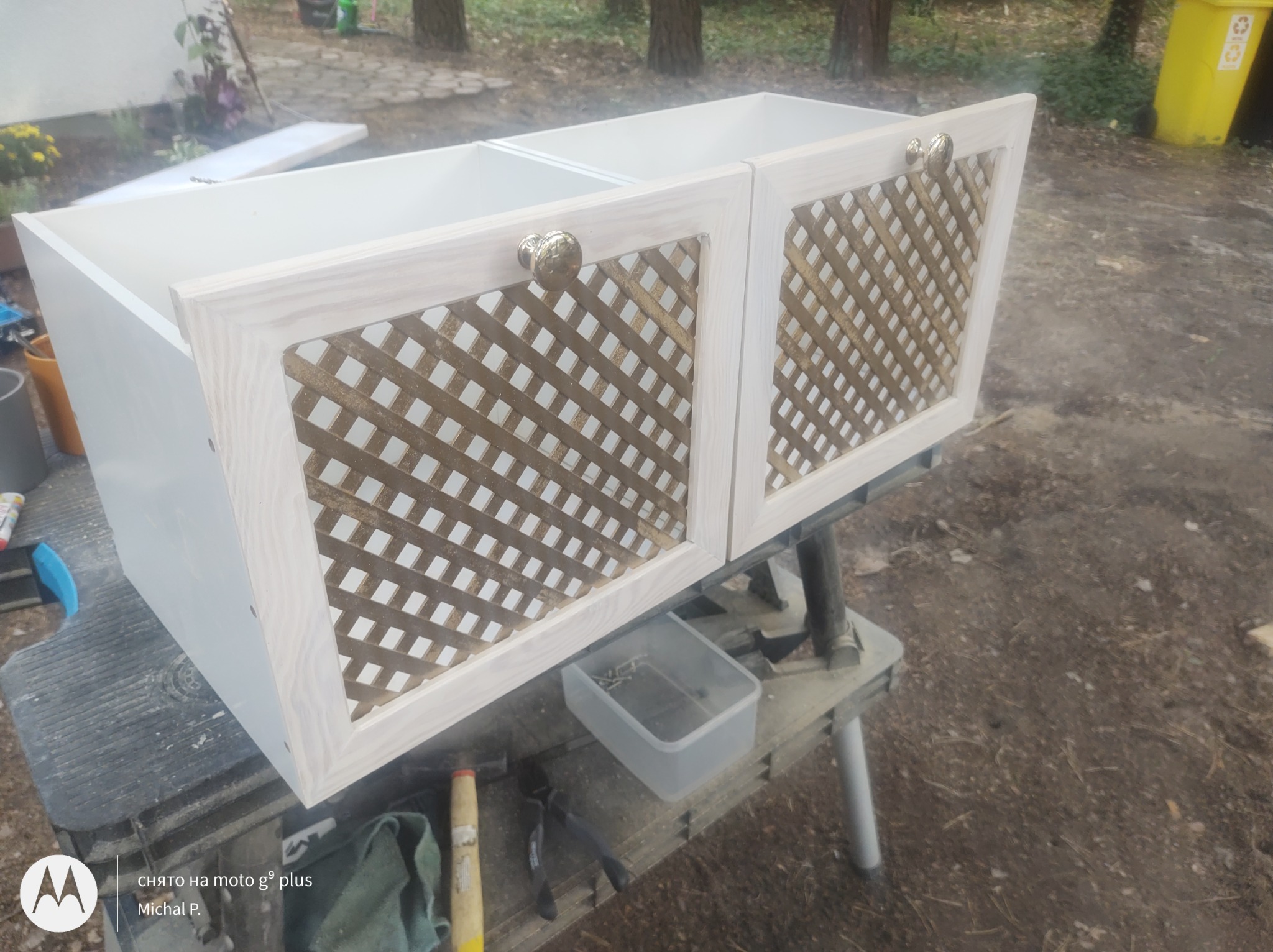 Dwa białe fronty szafek z ozdobną, ażurową kratką w kolorze złota i okrągłymi, metalowymi uchwytami, ustawione na warsztatowym stole, widoczne narzędzia i pudełko ze śrubami.