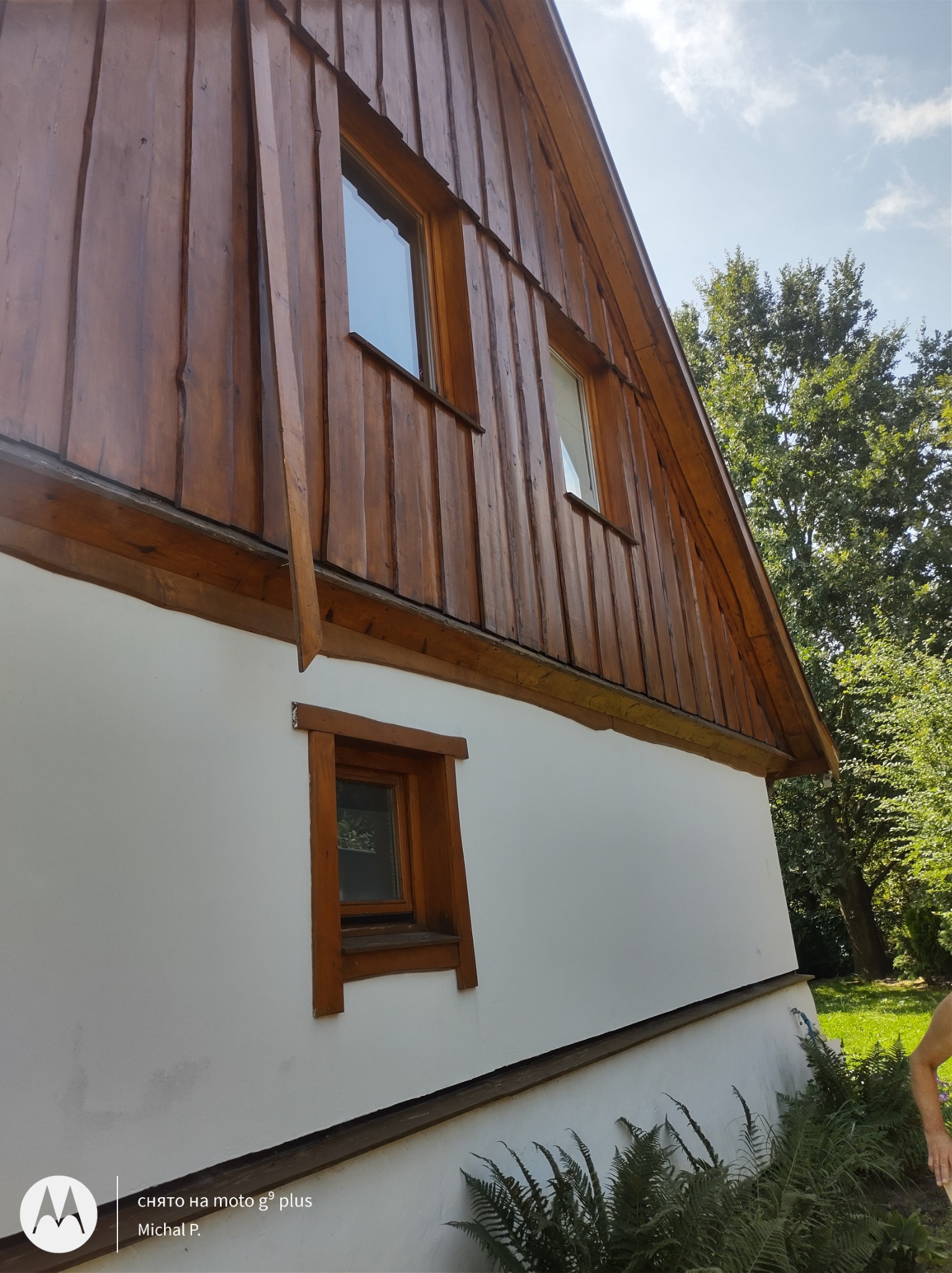 Fragment ściany domu z drewnianą elewacją i oknami z drewnianymi ramami, widok z dołu na tle zieleni.