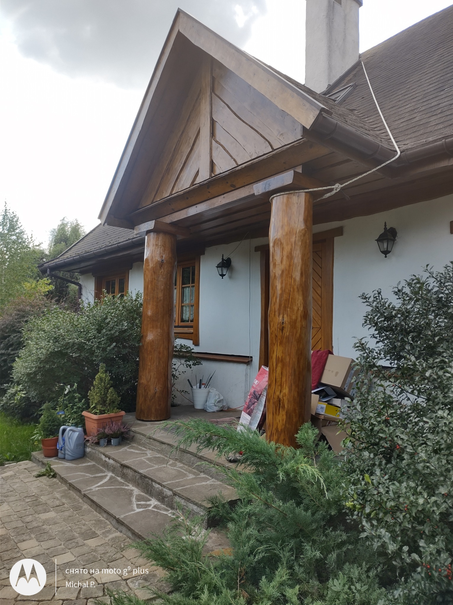 Wejście do domu z drewnianym zadaszeniem wspartym na okrągłych, drewnianych kolumnach, widoczne schody z kamiennych płyt i roślinność.