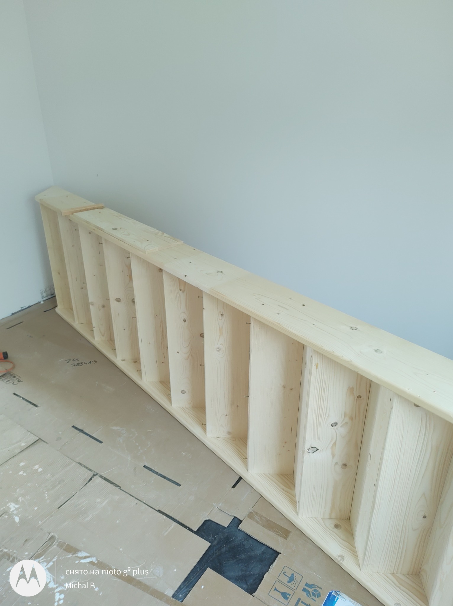 Surowa, drewniana konstrukcja antresoli, leżąca na podłodze zabezpieczonej kartonem, widoczne ślady ołówka i otwory po wkrętach, gotowa do montażu w jasnym wnętrzu.