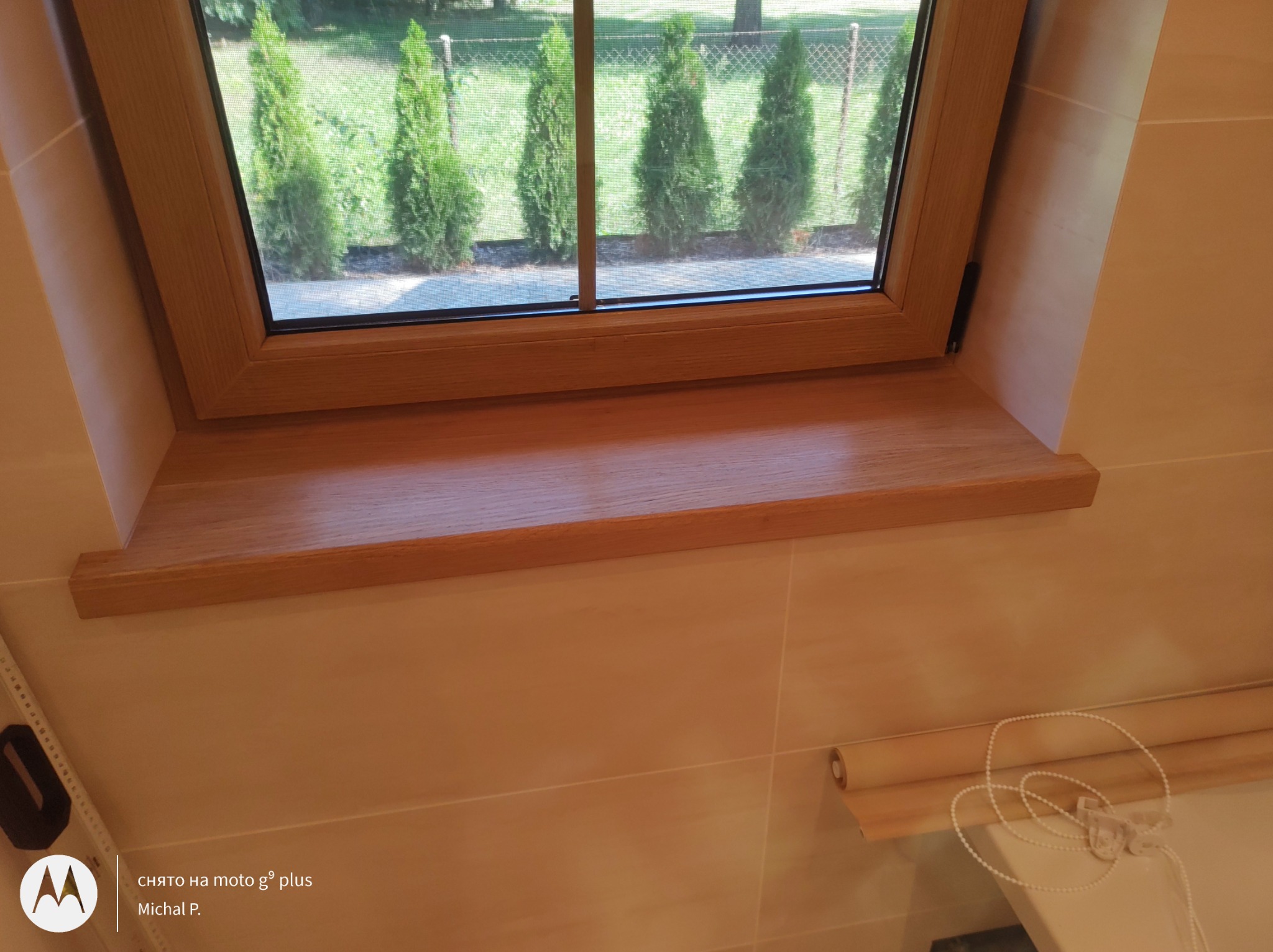 Drewniany parapet okienny z ramą okienną w kolorze naturalnego drewna, widok na rząd zielonych drzew iglastych za oknem, beżowe płytki na ścianie i zwinięta roleta okienna.