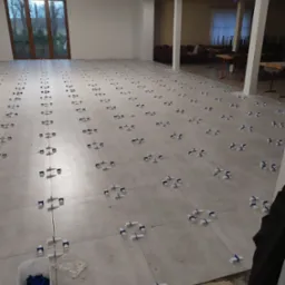 Układanie jasnoszarych płytek podłogowych z użyciem systemu poziomowania płytek (klipsy i kliny) w przestronnym, jasnym pomieszczeniu z widocznymi śladami kleju i wiaderkiem z klinami.