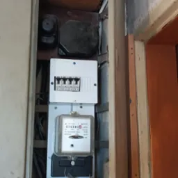 Otwarta skrzynka z licznikiem elektrycznym i bezpiecznikami wewnątrz, widoczne wystające przewody i uszkodzony tynk na ścianie.