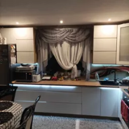 Nowoczesna kuchnia z białymi szafkami, drewnianym blatem, oświetleniem LED pod szafkami, szarymi zasłonami w oknie i dywanem shaggy. Widoczny fragment stołu z krzesłem.