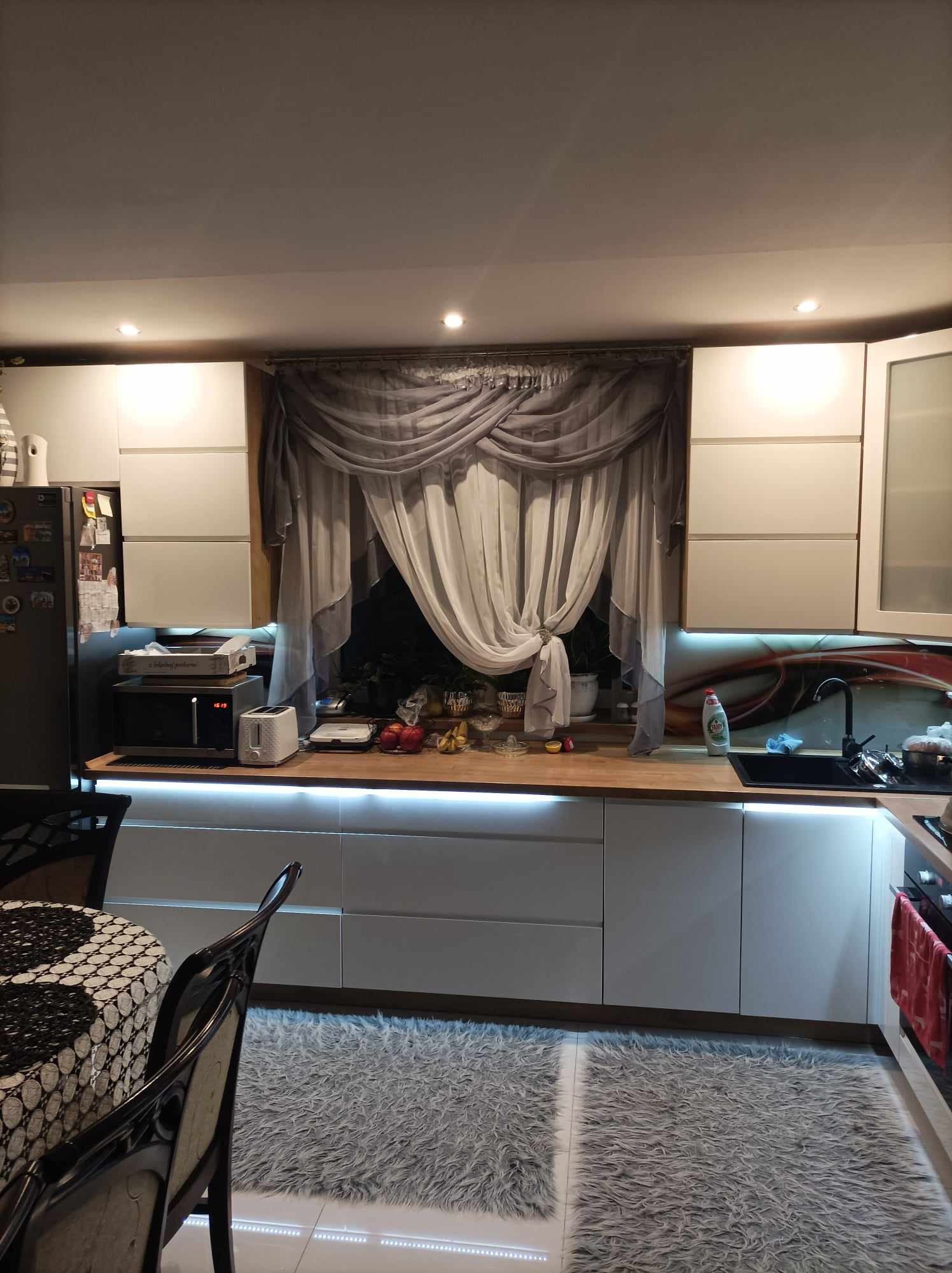 Nowoczesna kuchnia z białymi szafkami, drewnianym blatem, oświetleniem LED pod szafkami, szarymi zasłonami w oknie i dywanem shaggy. Widoczny fragment stołu z krzesłem.
