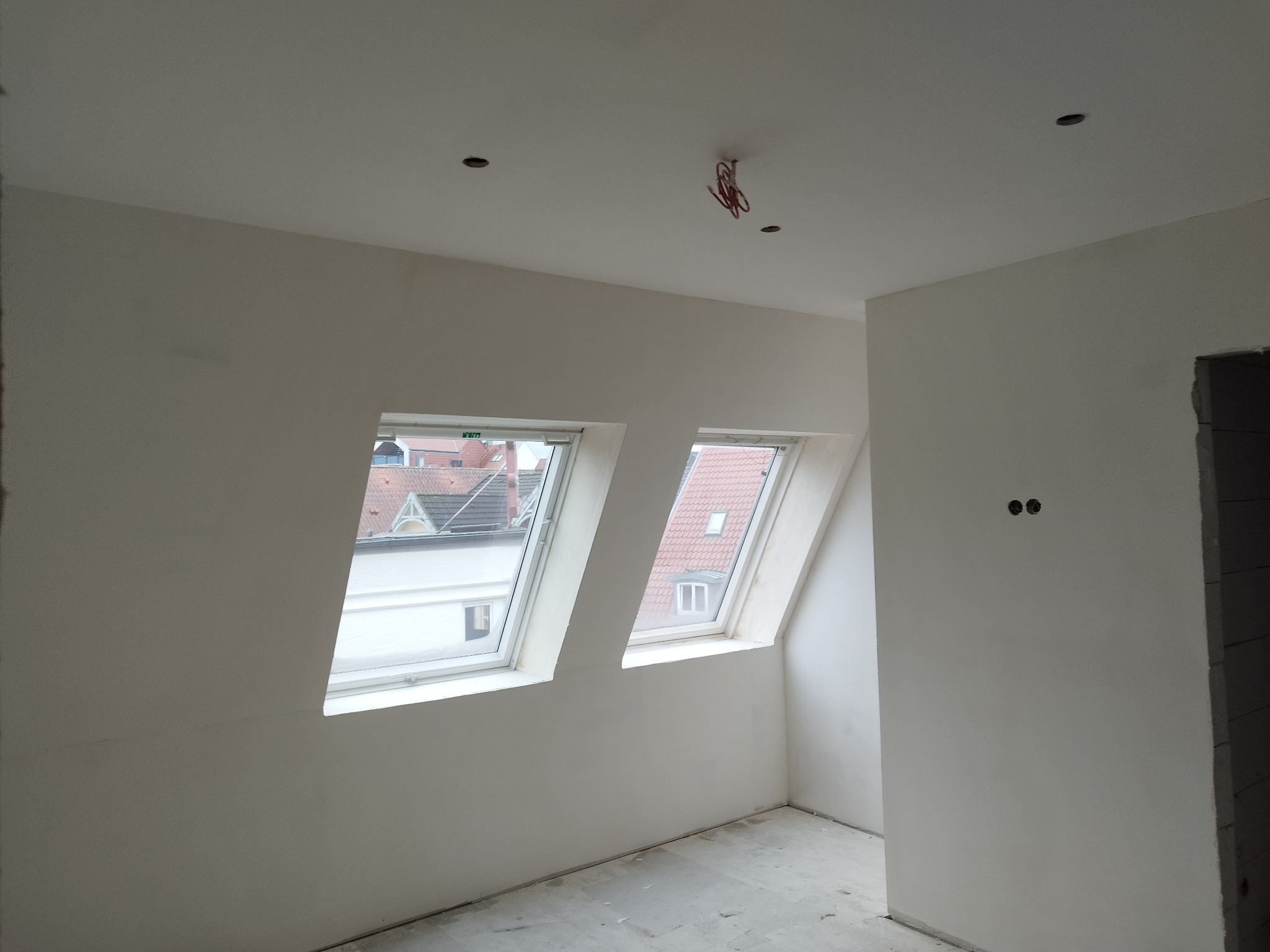 Wnętrze poddasza w Wodzisławiu Śląskim po szpachlowaniu ścian i sufitu, widoczne dwa okna dachowe i wystające kable oświetleniowe.