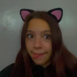 Portret młodej kobiety z brązowymi włosami, noszącej czarne opaski z r&oacute;żowymi kocimi uszami, robiącej głupią minę z wyciągniętym językiem i zezem na tle szarej ściany.