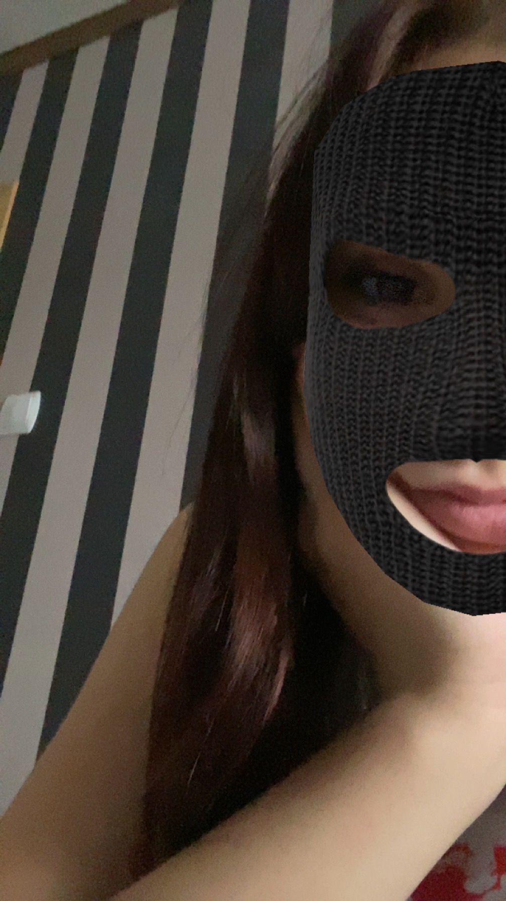 Osoba w czarnej kominiarce na tle ściany w czarno-białe pasy, widoczny fragment ramienia i włosów.