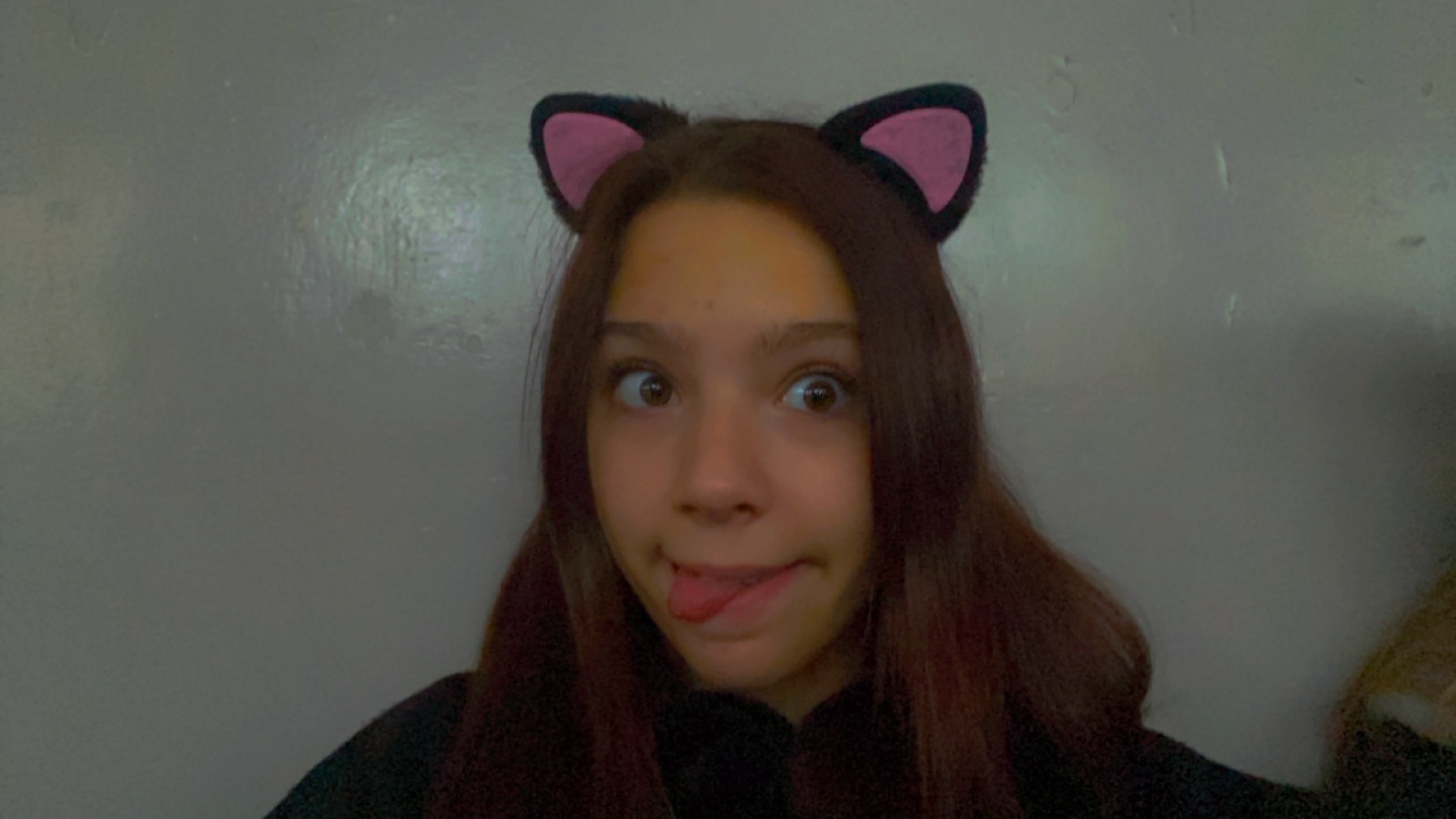 Portret młodej kobiety z brązowymi włosami, noszącej czarne opaski z różowymi kocimi uszami, robiącej głupią minę z wyciągniętym językiem i zezem na tle szarej ściany.