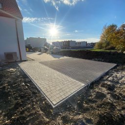 MEDIA GROUP SPÓŁKA Z OGRANICZONĄ ODPOWIEDZIALNOŚCIĄ - Przygotowany teren pod parking z częściowo ułożoną kostką brukową w jasnoszarym odcieniu i widoczną warstwą drenażową, w tle budynki mieszkalne i błękitne niebo z promieniami słońca.