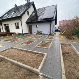 MEDIA GROUP SPÓŁKA Z OGRANICZONĄ ODPOWIEDZIALNOŚCIĄ - Układanie ciemnoszarej kostki brukowej na ścieżce w ogrodzie, z przygotowanymi rabatami ziemnymi, na tle domu z panelami słonecznymi i anteną.