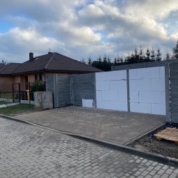 MEDIA GROUP SPÓŁKA Z OGRANICZONĄ ODPOWIEDZIALNOŚCIĄ - Ułożona szara kostka brukowa przed budynkiem z częściowo wzniesionym ogrodzeniem z szarych betonowych elementów i białych bloczków styropianowych, widoczne drzewa w tle.