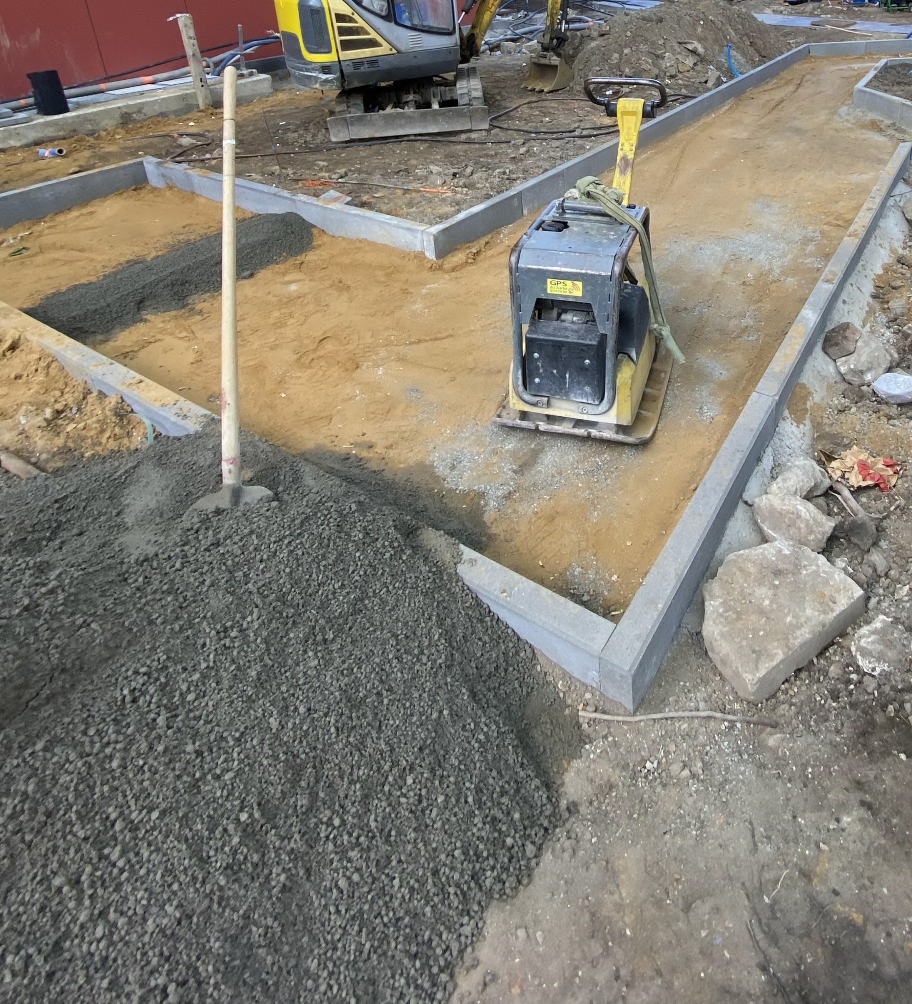 Przygotowanie podbudowy pod kostkę: ubijarka wibrująca na piasku, wykop z podsypką i stertą kruszywa, widoczne krawężniki i minikoparka w tle.