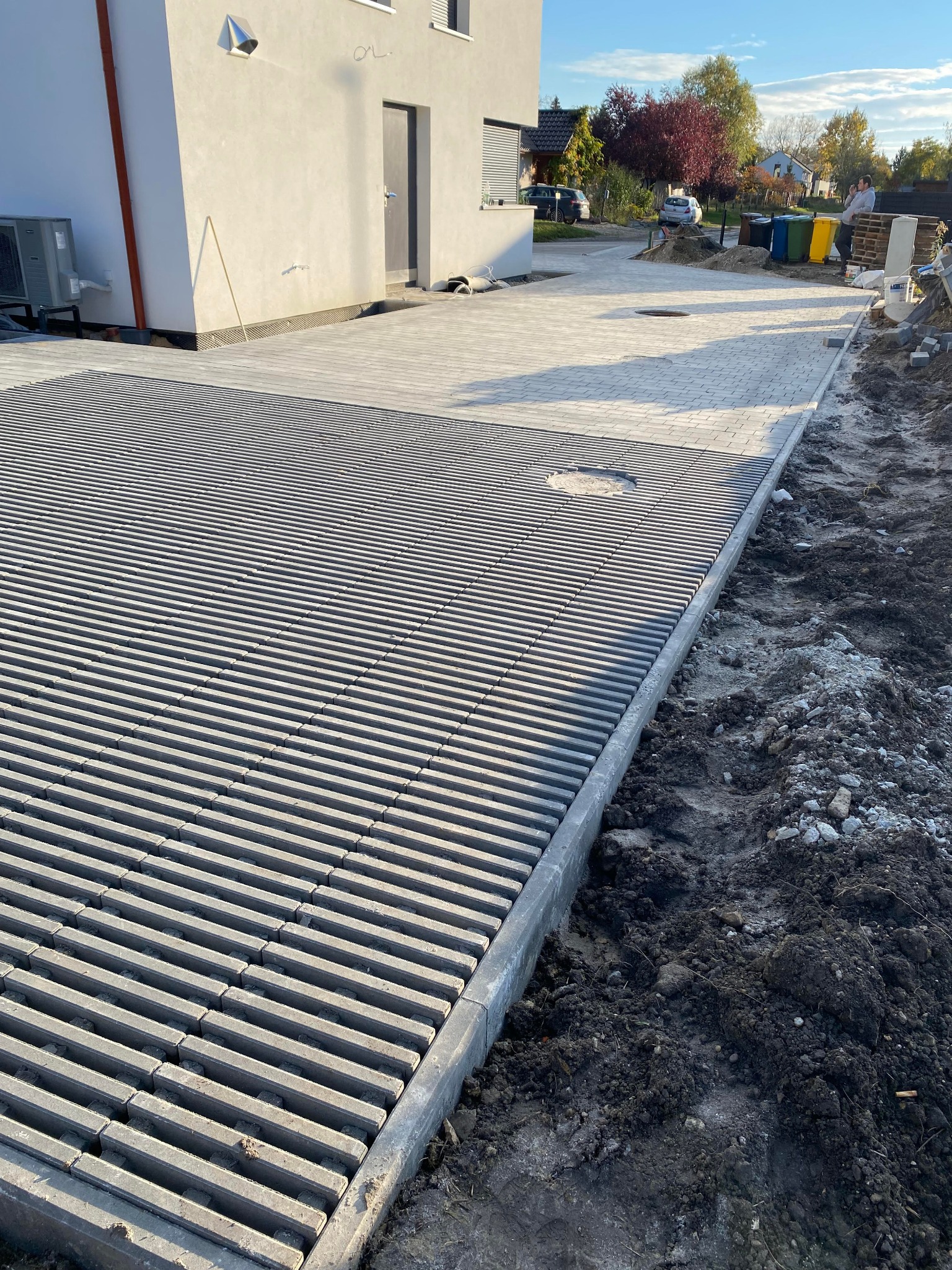 Ułożona, szara nawierzchnia parkingowa z ażurowych elementów betonowych, widoczna studzienka kanalizacyjna, obrzeża z kostki, w tle budynek mieszkalny i przygotowany teren pod dalsze prace brukarskie.