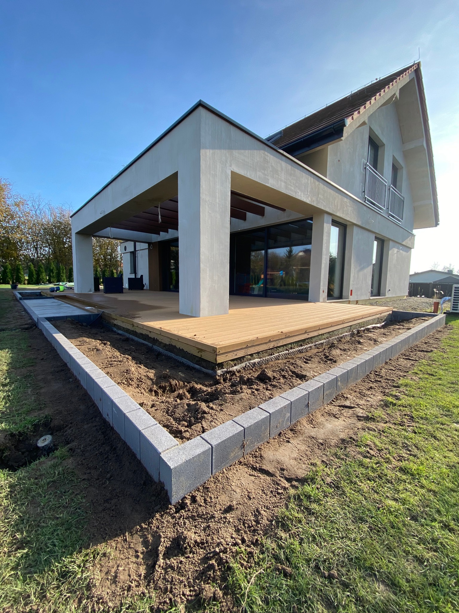 Nowoczesny dom z drewnianym tarasem i obrzeżem z szarej kostki palisadowej otaczającym rabatę z ziemią, widok w słoneczny dzień.