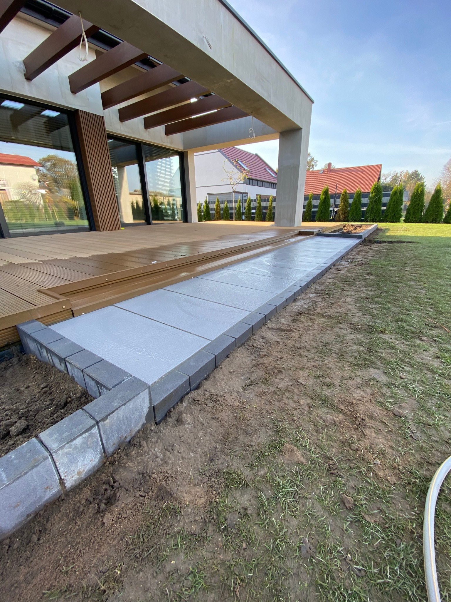 Świeżo ułożony chodnik z jasnoszarych płyt obramowany ciemniejszą kostką, prowadzący od drewnianego tarasu nowoczesnego domu w kierunku trawnika.