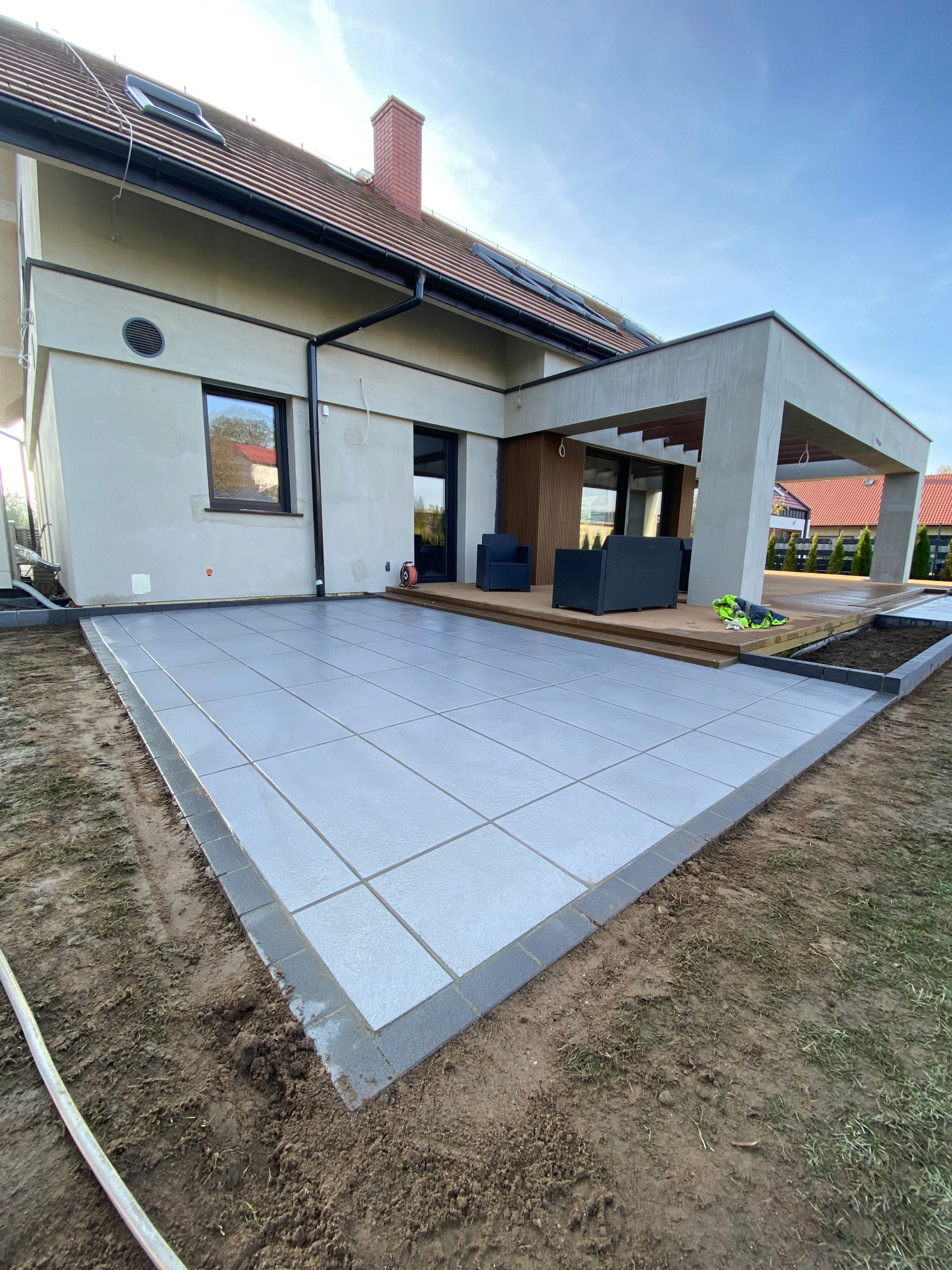 Nowoczesny taras z szarych płyt betonowych obramowany ciemniejszą kostką, przylegający do domu z drewnianym zadaszeniem i meblami ogrodowymi w tle, świeżo po zakończeniu prac.