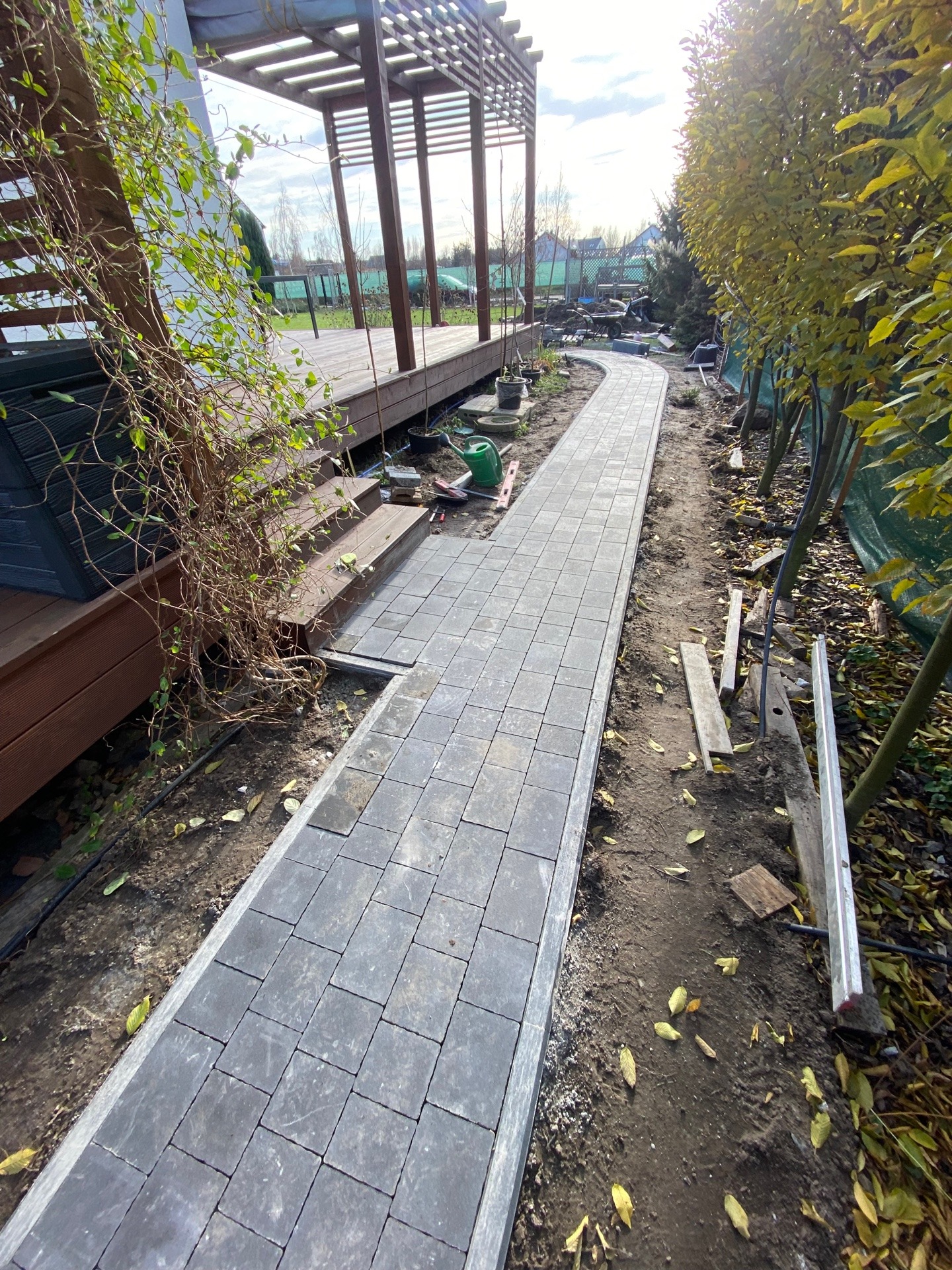 Ścieżka z szarej kostki brukowej w trakcie układania, widoczna obok drewnianego tarasu z pergolą, z narzędziami i zielonymi roślinami w tle.
