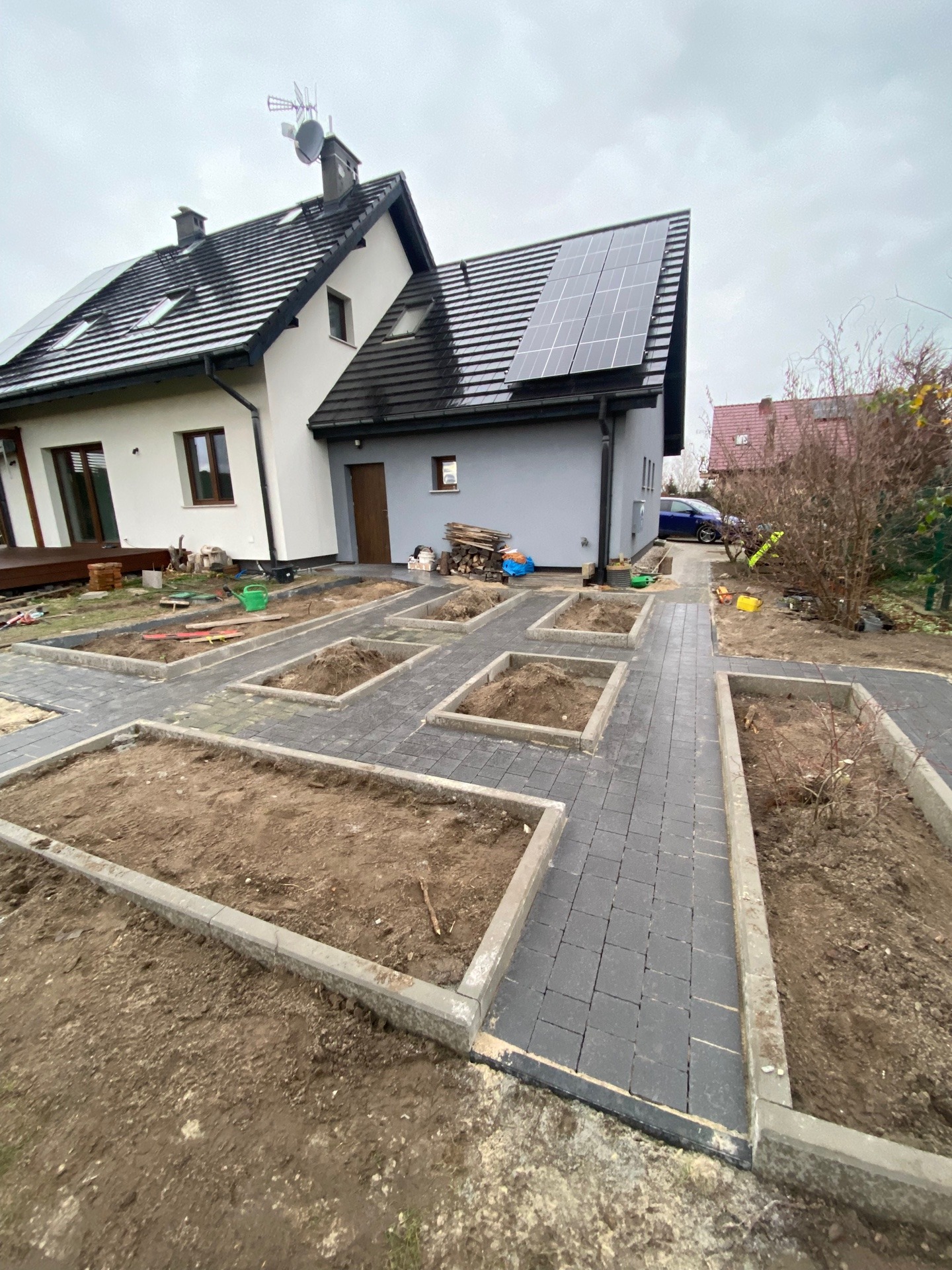 Układanie ciemnoszarej kostki brukowej na ścieżce w ogrodzie, z przygotowanymi rabatami ziemnymi, na tle domu z panelami słonecznymi i anteną.