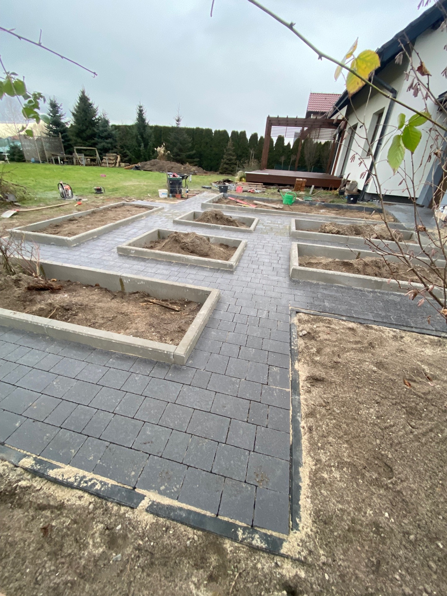 Układanie ciemnoszarej kostki brukowej z obrzeżami tworzącymi rabaty na ziemię, widoczne narzędzia ogrodnicze i trawnik w tle, pochmurne niebo.