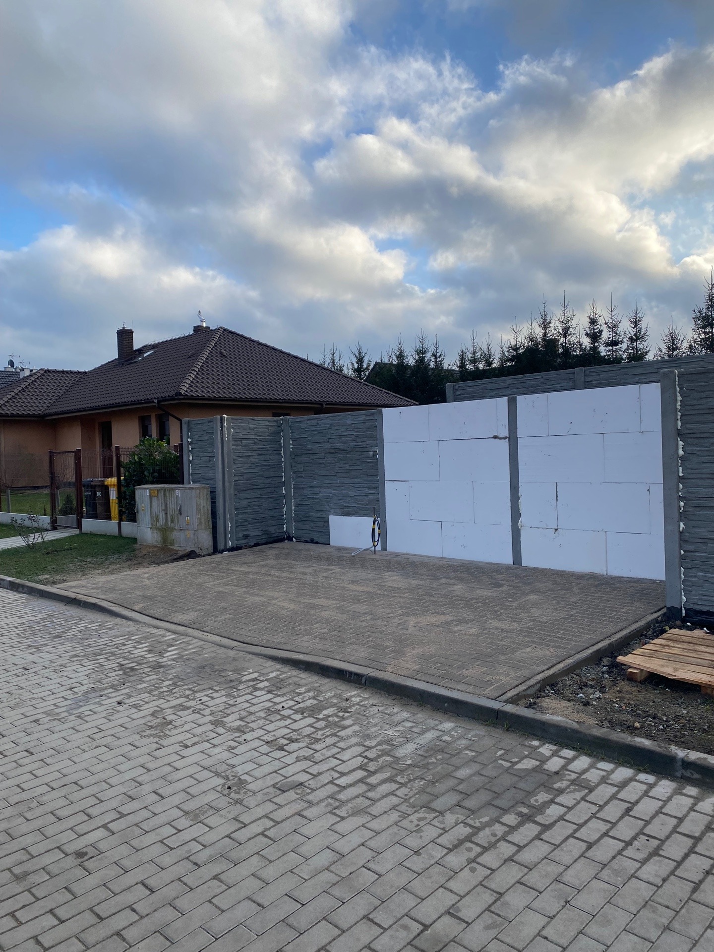 Ułożona szara kostka brukowa przed budynkiem z częściowo wzniesionym ogrodzeniem z szarych betonowych elementów i białych bloczków styropianowych, widoczne drzewa w tle.