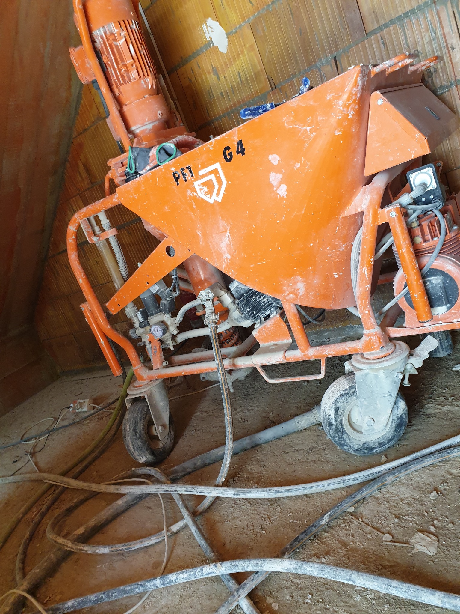 Pomarańczowa maszyna do tynków maszynowych PFT G4, widoczna pod kątem, z przewodami leżącymi na ziemi, w surowym wnętrzu z ceglanym murem.
