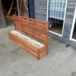 Brązowa, drewniana skrzynia na kwiaty z ażurowym panelem na tle betonowego podłoża, drzwi stodoły i okna.