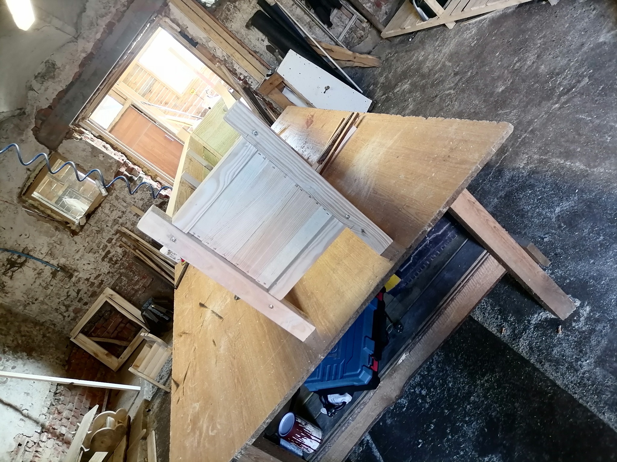 Warsztat stolarski z surowym drewnem, narzędziami i niedokończonym projektem meblowym na drewnianym stole roboczym w pomieszczeniu z oknem.