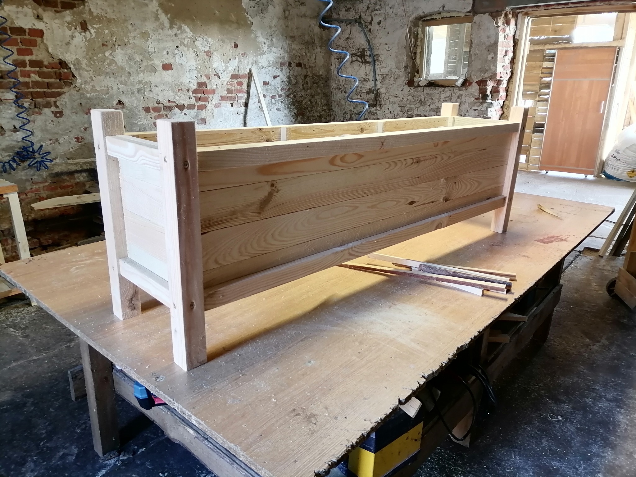 Surowa drewniana skrzynia na nogach, w trakcie budowy, umieszczona na roboczym stole w warsztacie z ceglaną ścianą w tle.