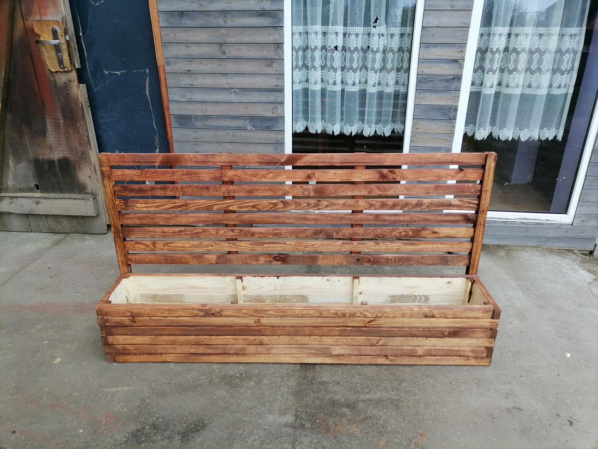 Drewniana skrzynia na kwiaty z oparciem, bejcowana na brązowo, na tle szarej ściany z oknem z firanką.