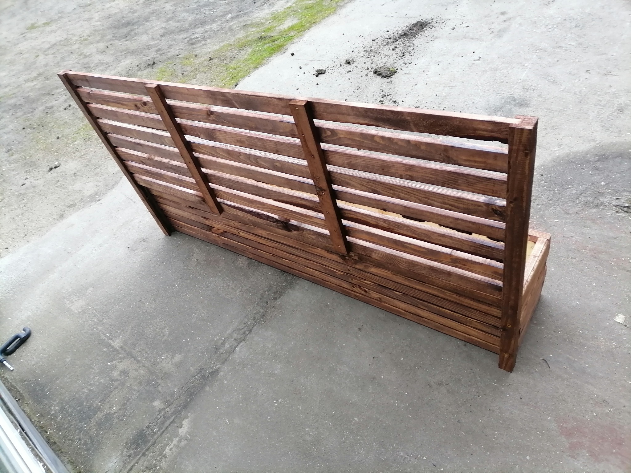 Drewniana konstrukcja z poziomych listew, prawdopodobnie element ogrodzenia lub osłony, impregnowana na ciemny brąz, widoczna na betonowym podłożu z narzędziem obok.