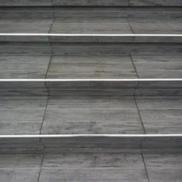 Schody wyłożone szarymi płytkami imitującymi drewno, z metalowymi listwami antypoślizgowymi na krawędziach stopni.