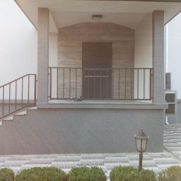 GSM SPOLKA Z OGRANICZONA ODPOWIEDZIALNOSCIA - Zadaszone wejście do domu z betonowymi schodami, metalową balustradą i elewacją z teksturą drewna, oświetlone lampą ogrodową.